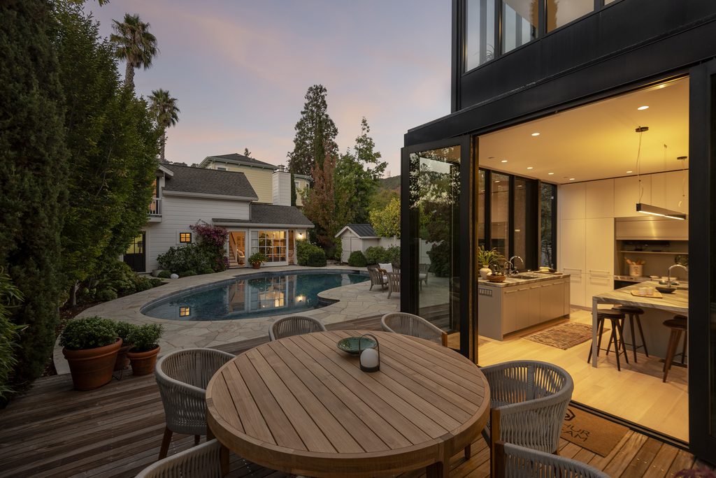 An indoor-outdoor kitchen open to a redwood deck, swimming pool, and an ADU/pool house in the twilight light.